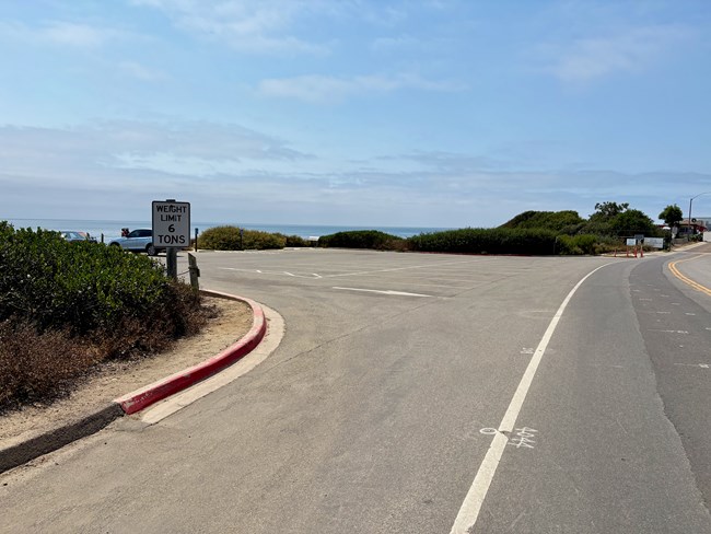 Empty parking lot by the ocean, weight limit sign, red curb, bushes, and a curving road.