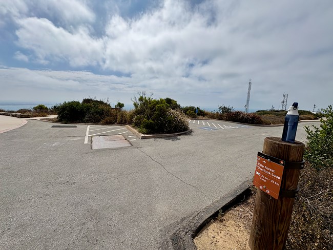 Empty parking lot, trail sign, water bottle on post, bushes, and radio towers under cloudy sky.