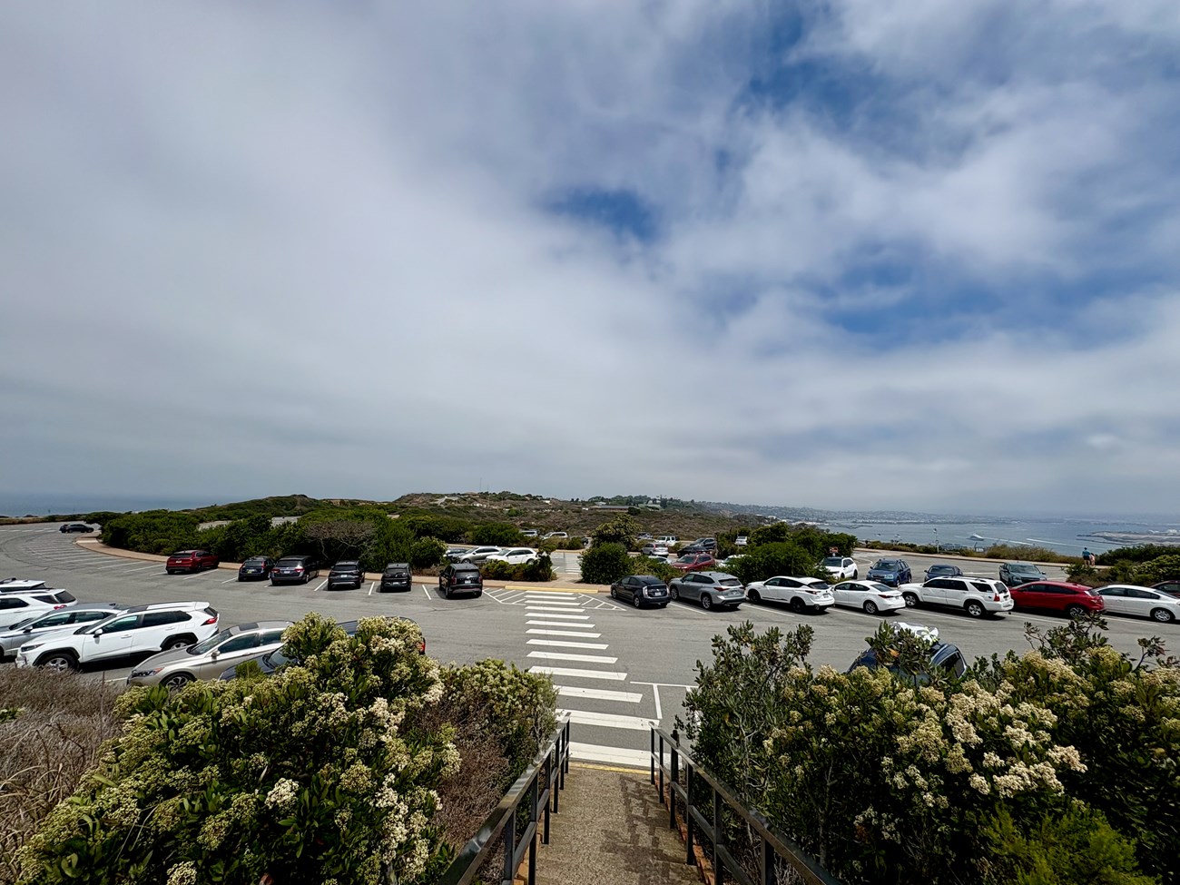 Coastal parking lot with many cars, bushes, and ocean visible under a cloudy sky.