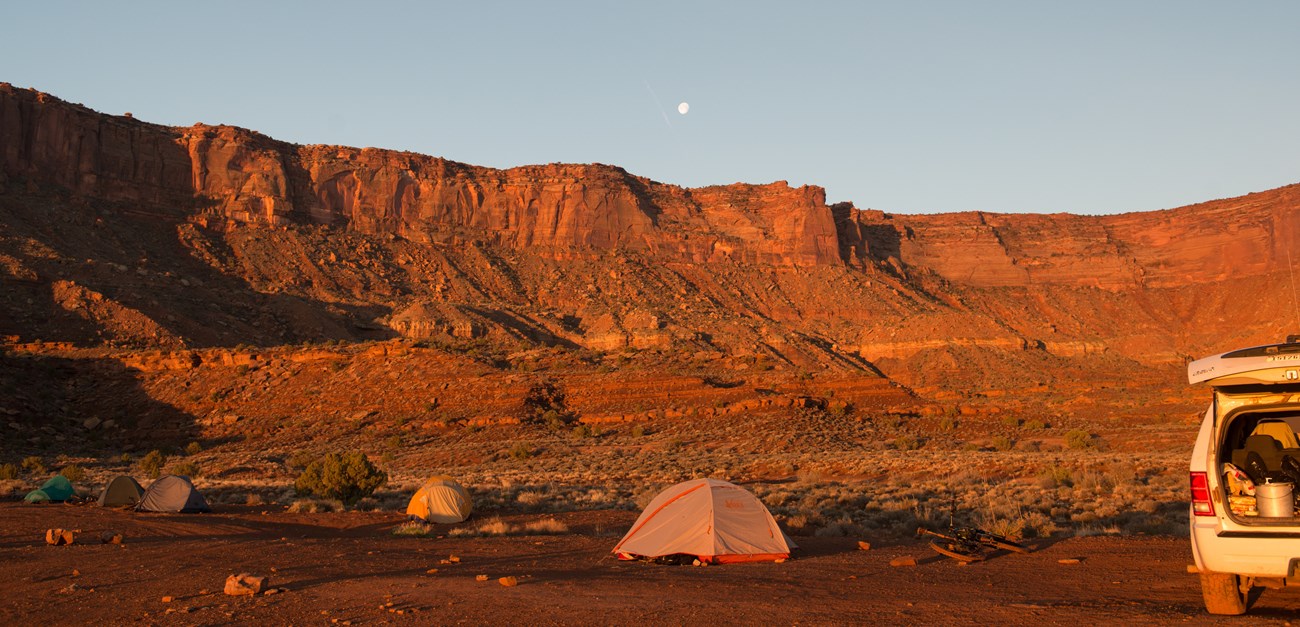 4 tents and a vehicle with the trunk open in a white rim campsite with sandstone clidds in the background.