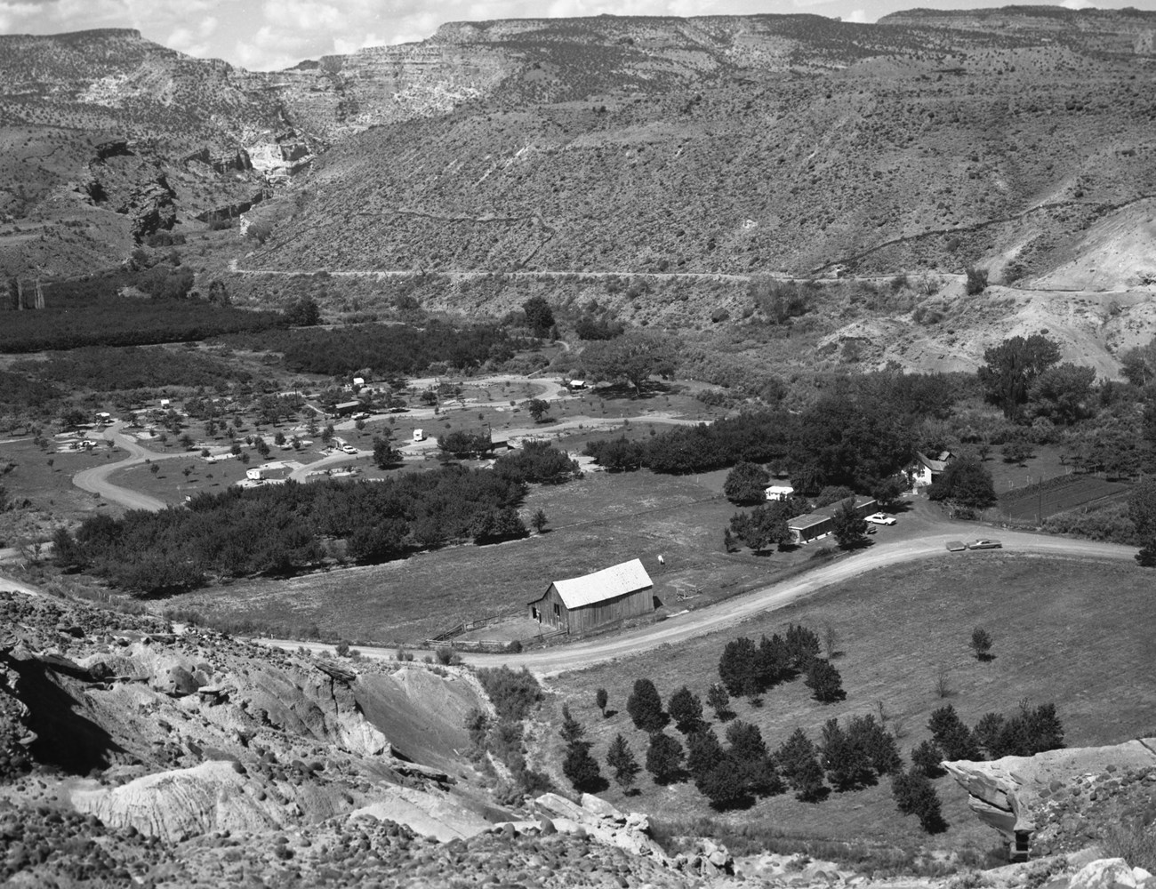 Black and white photo of large wooden barn, small house, low-slung one-story building alongside a road, with trees, campgrounds, rocky cliffs, and sky in background.