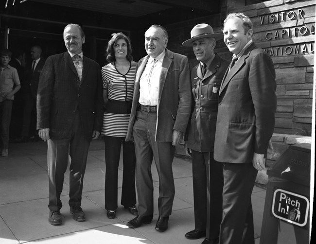 Black and white photo of four men and one woman standing in front of stone building with Visitor/ Capitol/ National on it.