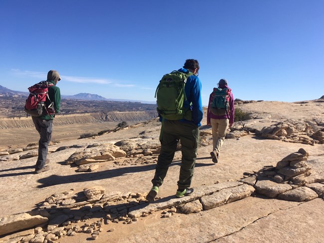 Three people walking on tan sandstone, with some sparse vegetation, and views into a wide valley and blue sky above.