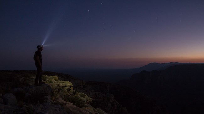 A hiker wearing a headlamp looks over the land as twilight sets in