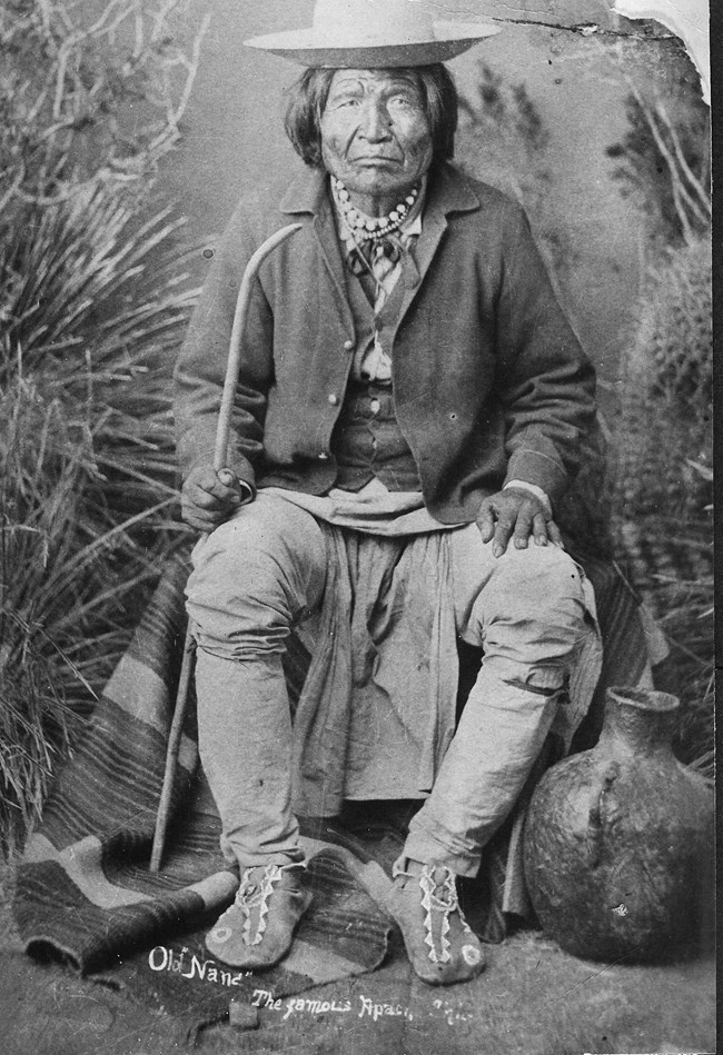 Black and white photo of an older man with short hair, wearing a jacket and a hat. He is seated in front of a striped blanket, holding a staff.