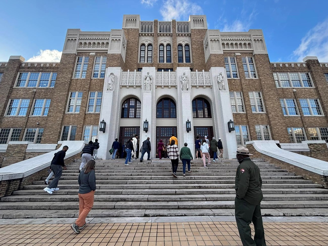 A group of people walk up the wide stone steps of Little Rock Central High School, a large brick and white stone building with tall windows and ornate carvings, as an NPS ranger in uniform looks on.
