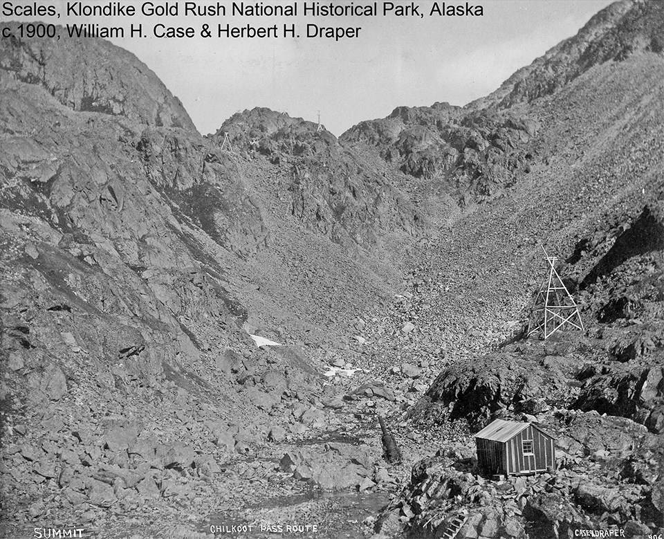 Black and white image of rocky mountain pass with one wooden building in foreground.