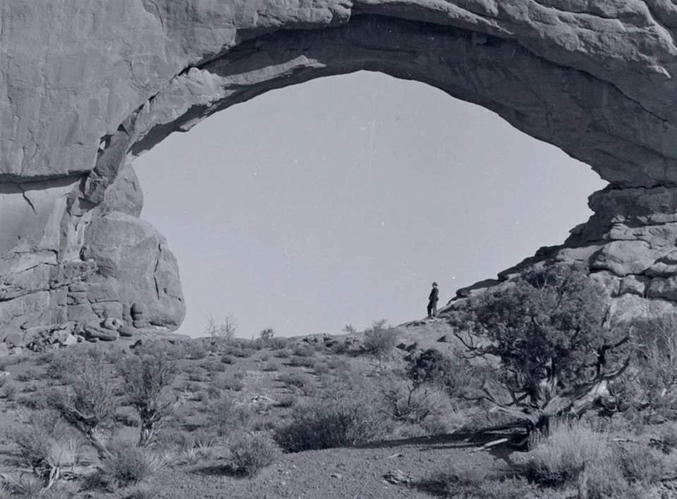 a black and white photo of North Window. A man stands under the arch on the right side. There are plants and soil on the stone below the arch.