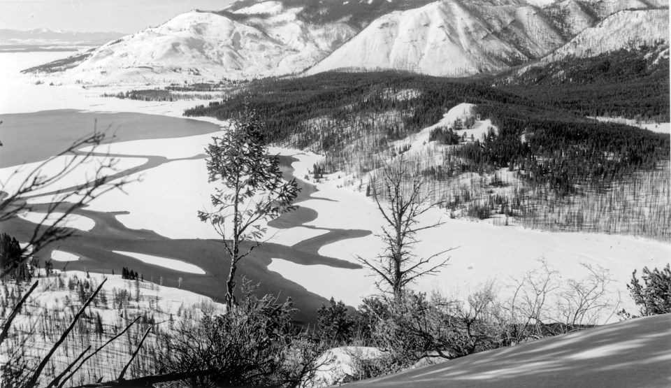 A female skier dressed in early 1900s garb stands in front of a snowy, mountainous, forested landscape above a lake.