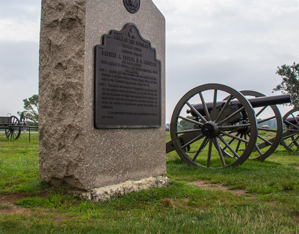 Four Civil War veterans sit at the base of a small monument as one of them points with his cane. A cannon sits in the background.