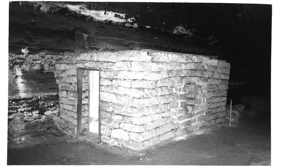 A group of people sit and stand around a rectangular stone hut inside a cave in 1912.