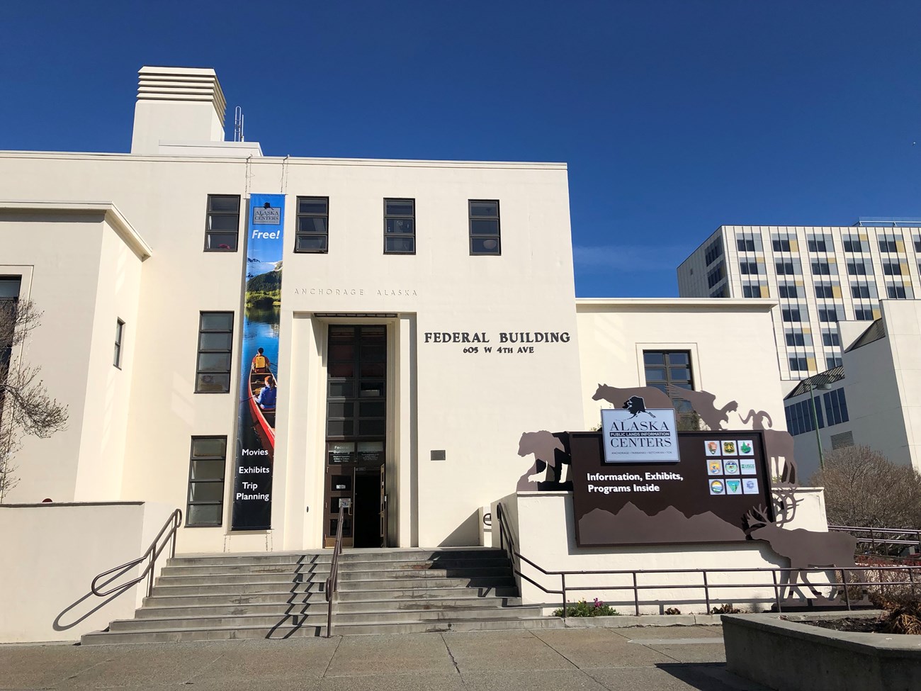 Outdoors; sandstone building with "Federal Building" on side as well as banner and separate sign inviting public inside.