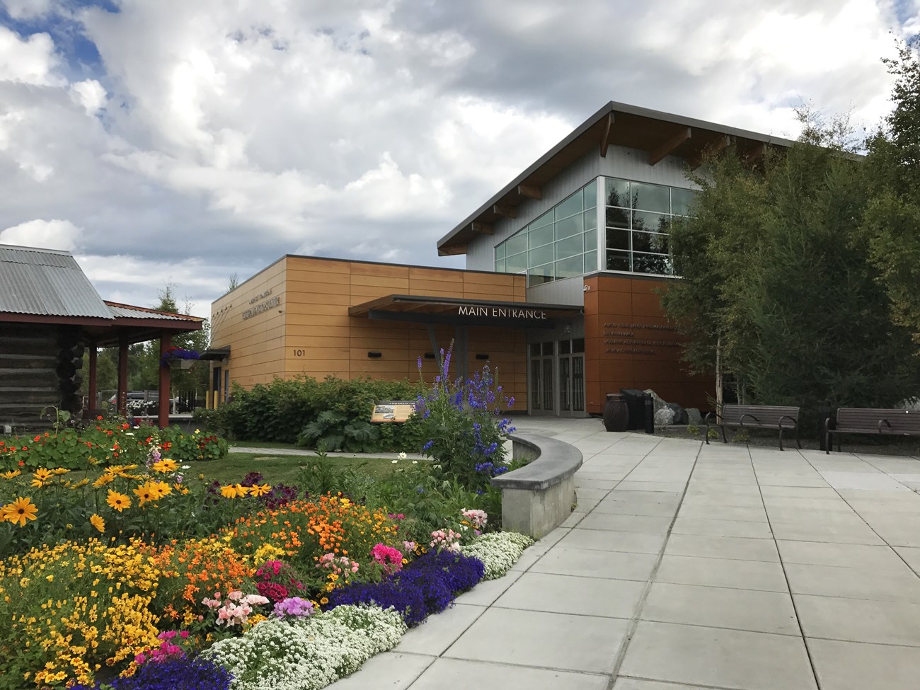 Outdoors; flower beds in forefront to left of cement sidewalk leadng to a large wooden block building.