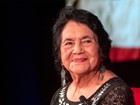 Older women in black shirt with white pattern and black hair sits with her hands in lap, smiling.