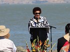 Black woman stands at podium speaking to crowd in front of river