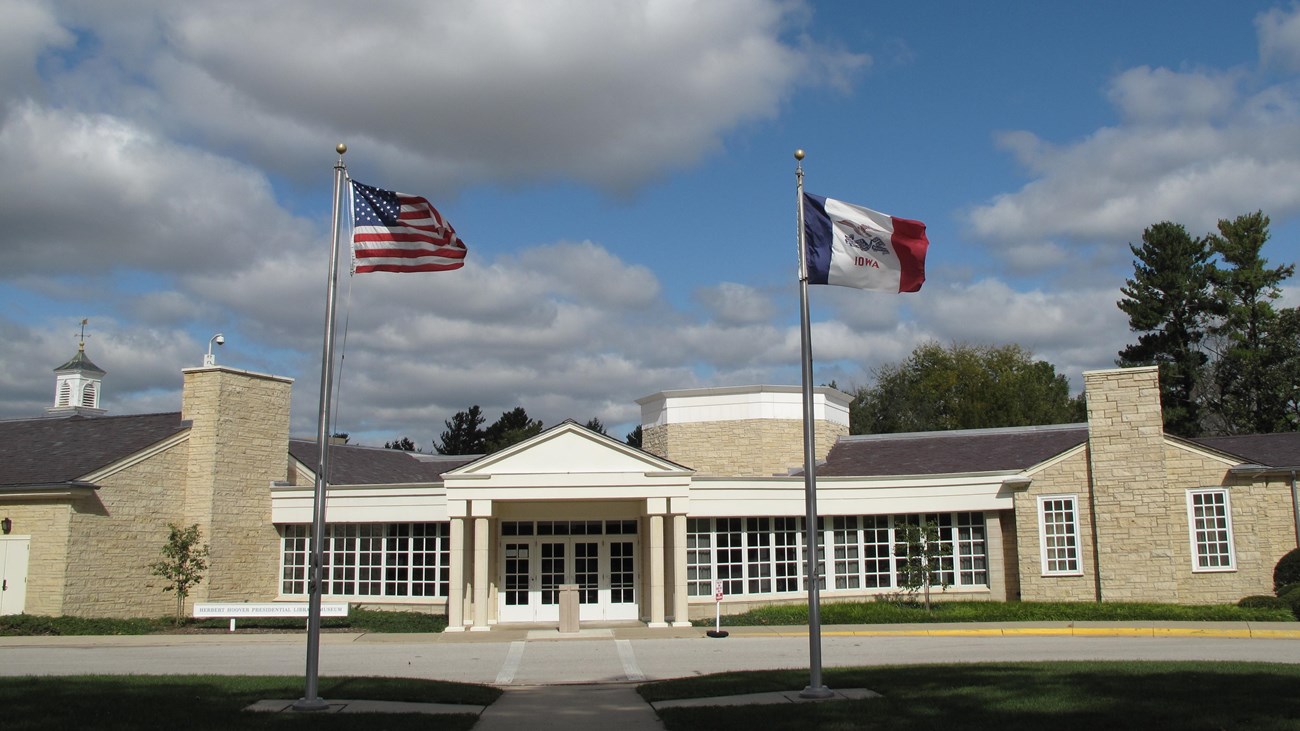 A sprawling one story public building of rough-faced yellowish stone has a white portico entrance.