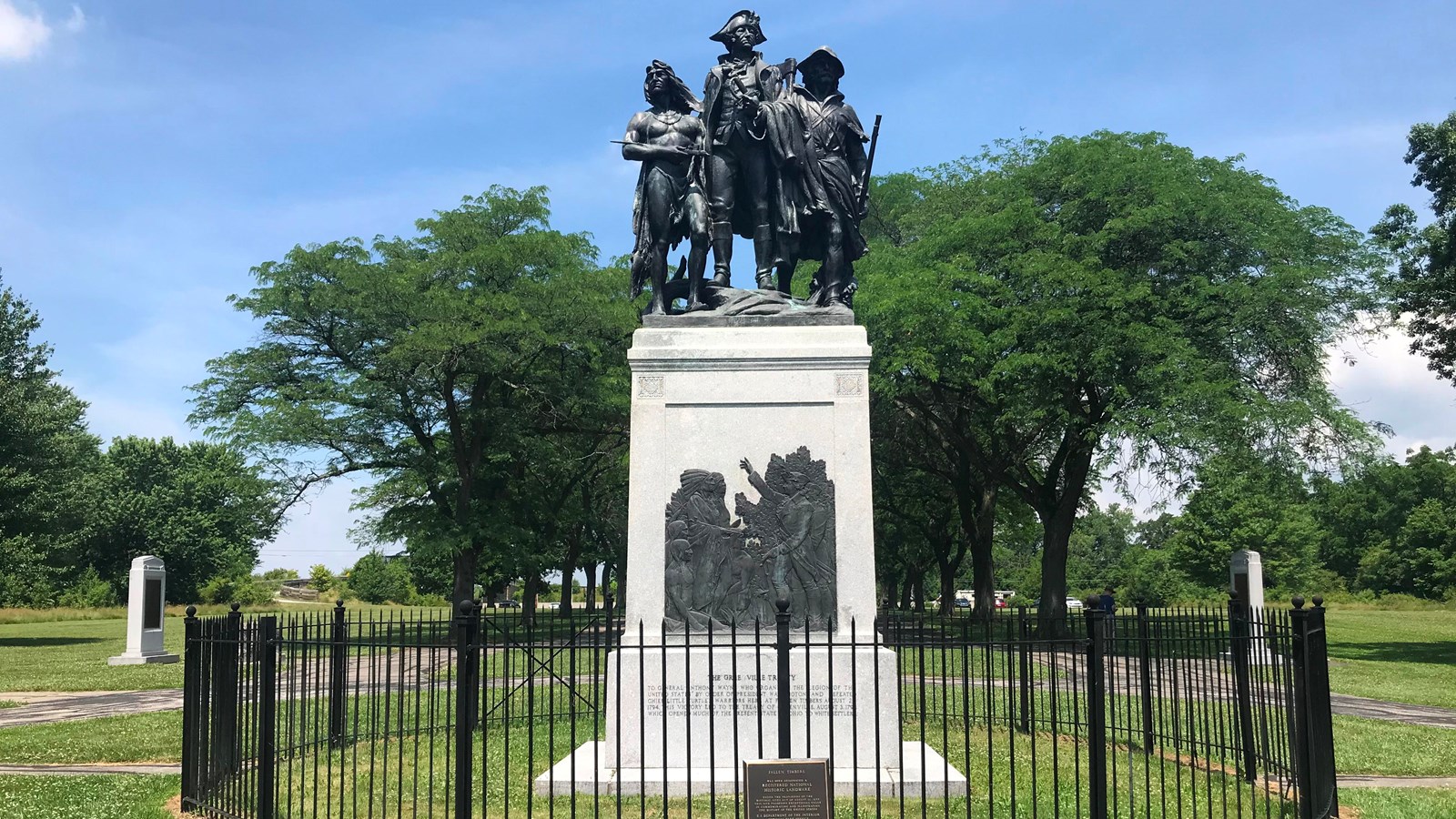 A path leads to a black fence surrounding a white pillar with a plaque and a statue of 3 men on top.