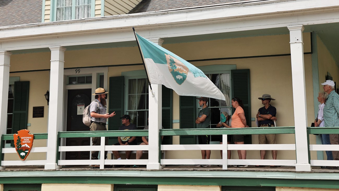 Park ranger talking un a covered porch of a cream house to several people. 