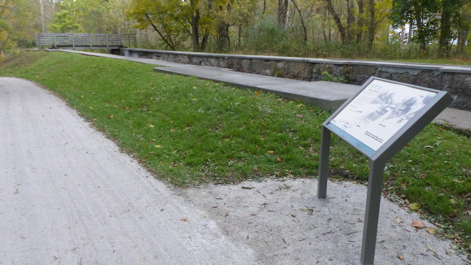 A trail to the left of a canal lock\'s cement walls; a wooden footbridge crosses over the far end.