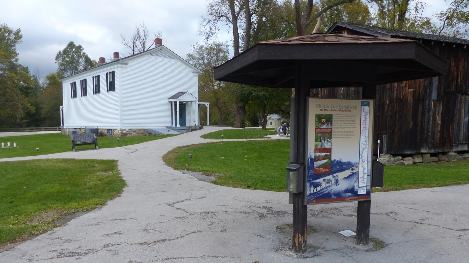 A branching walkway leads from a three sided kiosk past a brown barn to a two story white building.