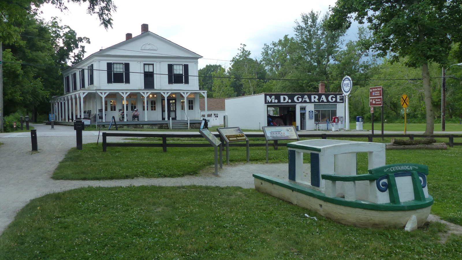 White cement play boat with green trim in grass beside three panels. M.D. Garage across the street.