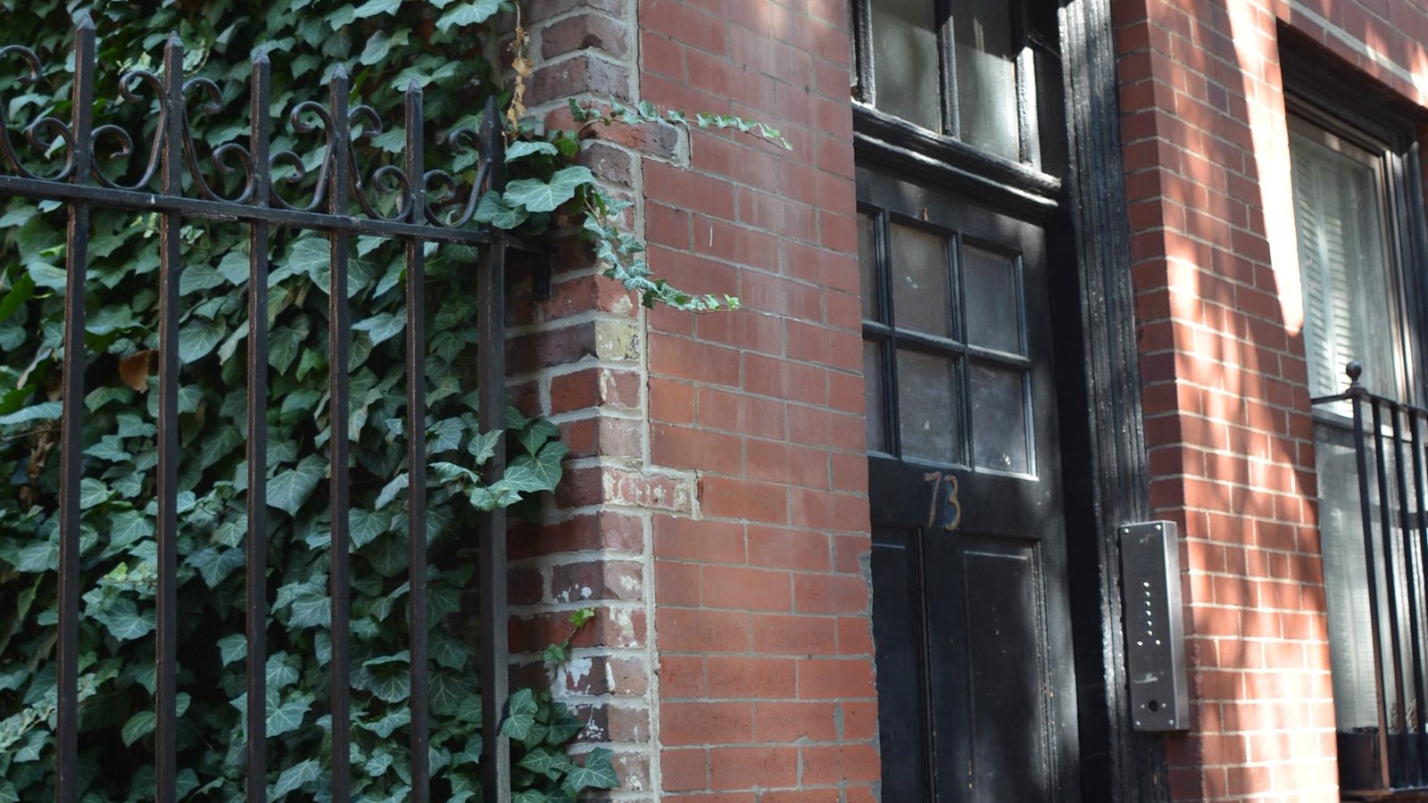 Red brick building with a black door on the right with a metal fence and bushes on the left.