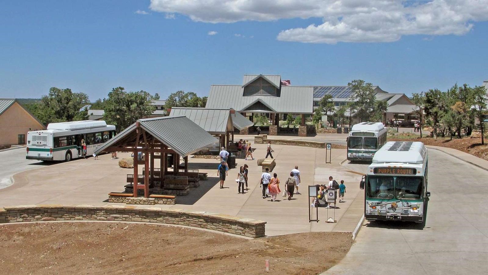 Three large white and green buses circle a transit center with signs and shade structures.