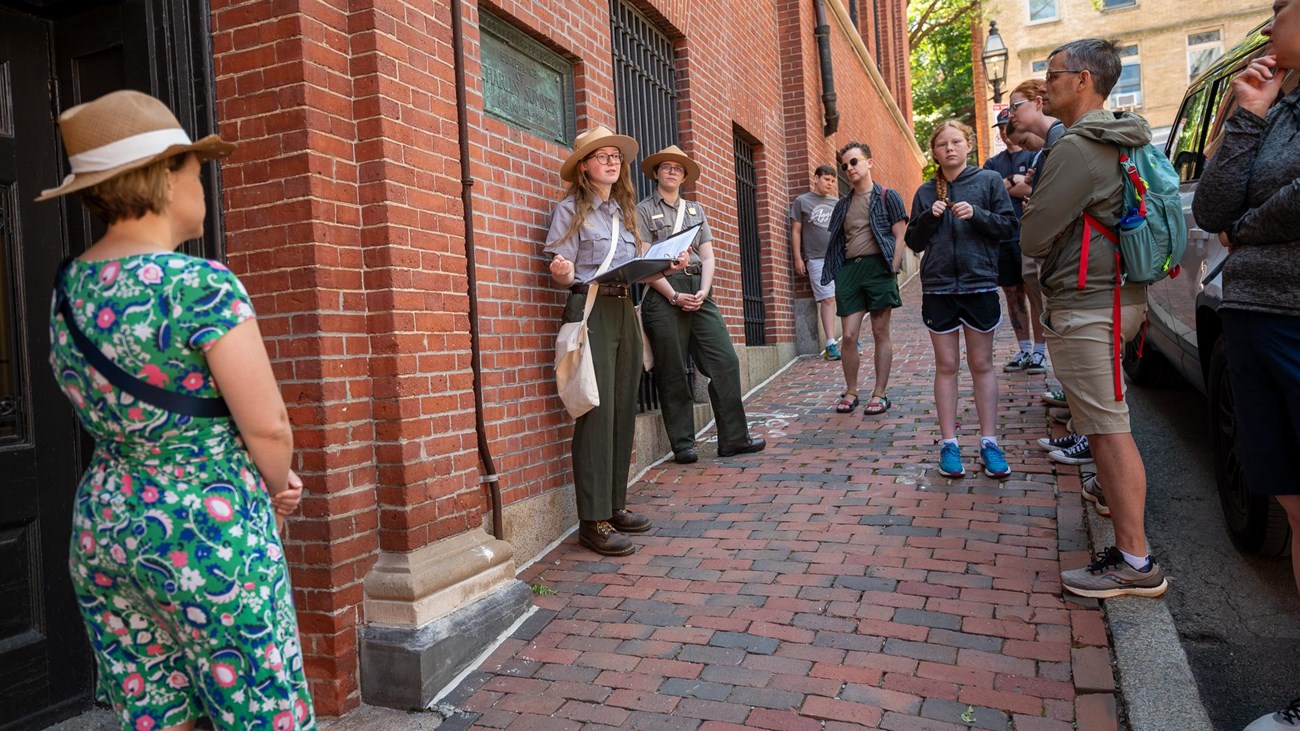 Two park rangers stand in front of a crowd talking next to a brick building