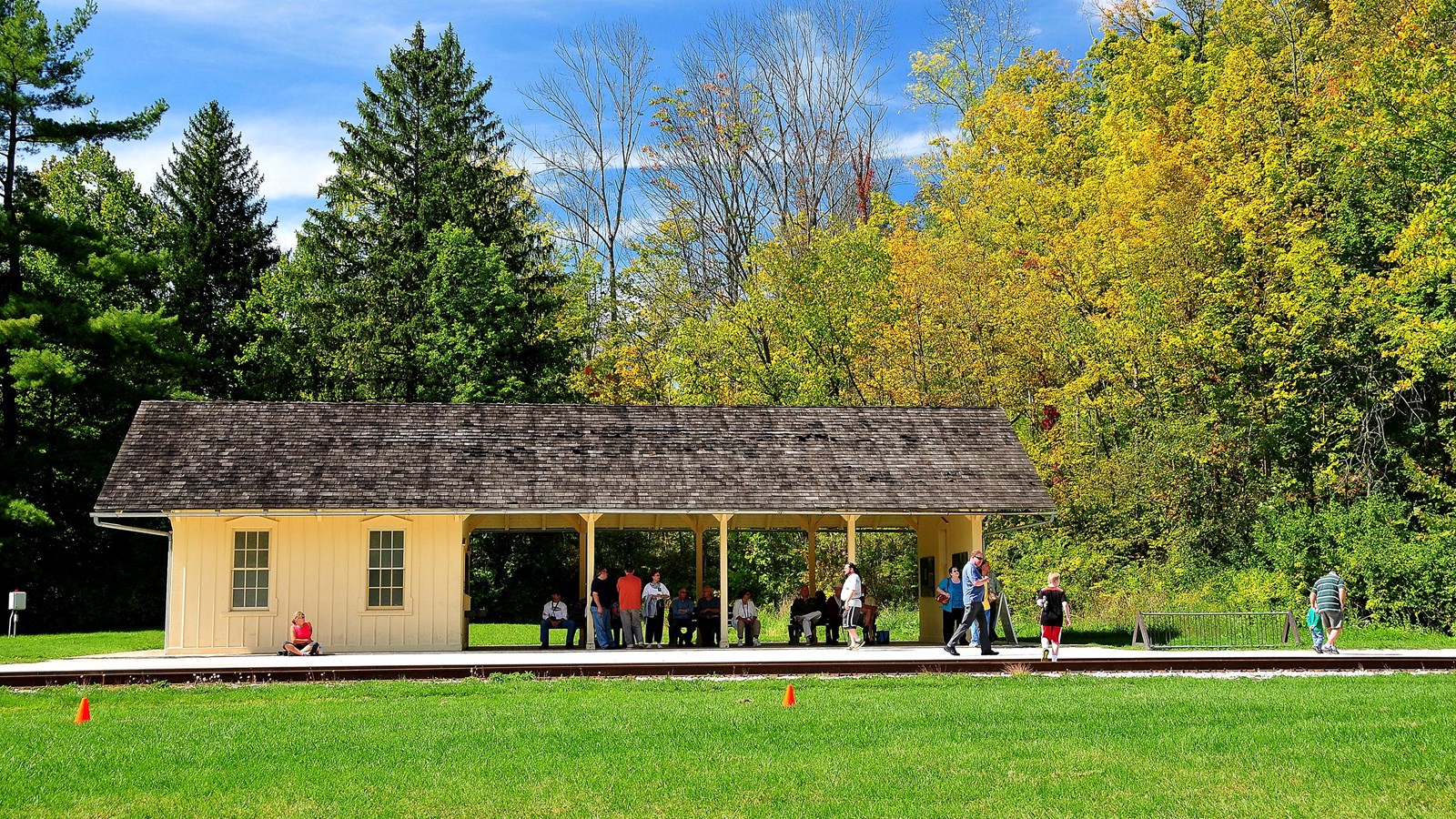 Beyond lawn and train tracks, people wait in a yellow station with open sides and enclosed left end.