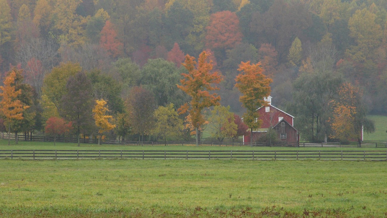 Scenic view of red barn with grassy fields in foreground and hillside of colorful leaves behind.
