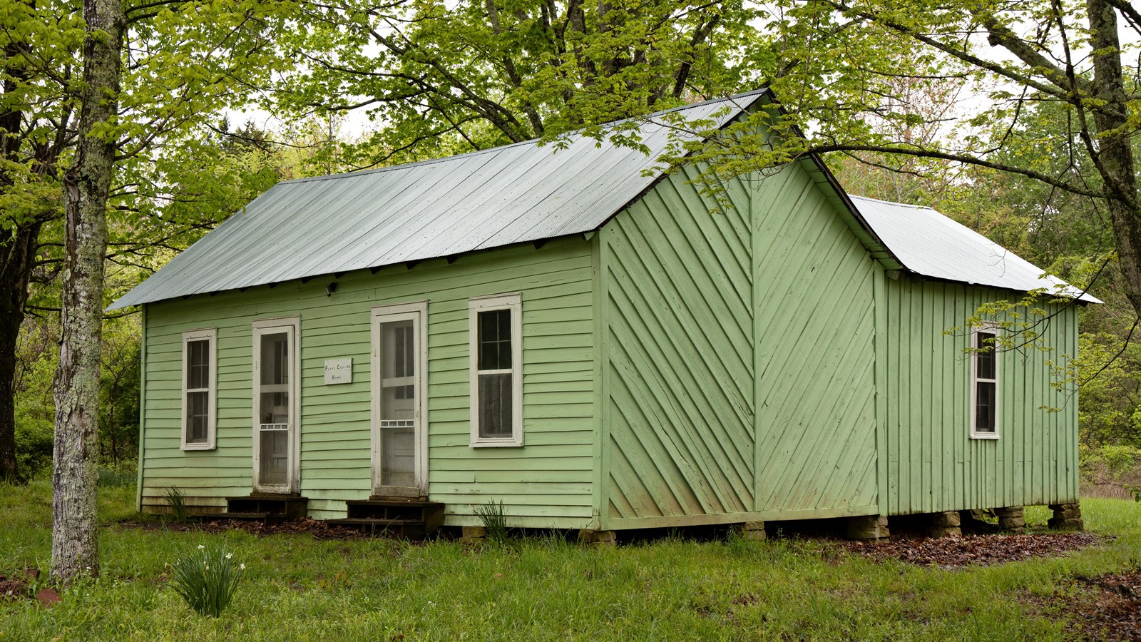 A simple wooden house with green paint and two front doors, sitting in a grassy field.