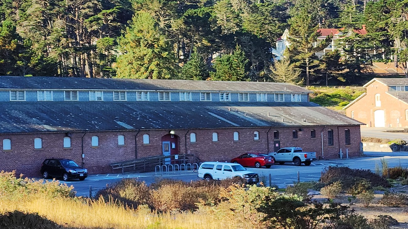 Long brick building with a parking lot in front surrounded by trees.