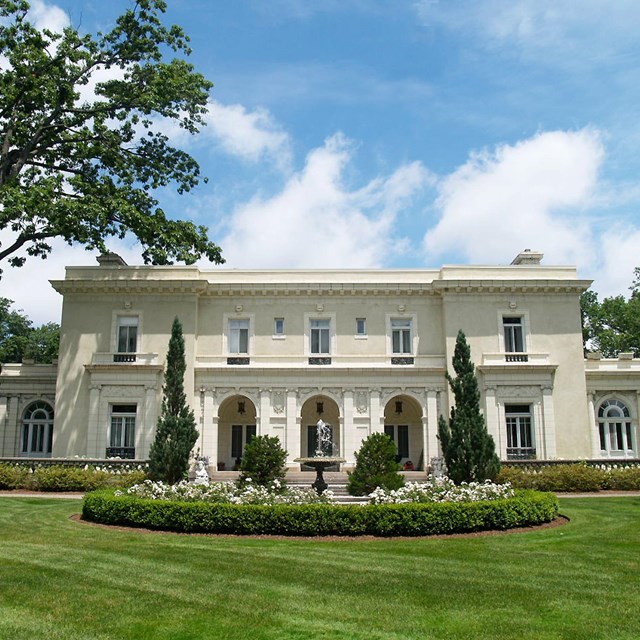 Large white ornate home with blue sky background and large grass lawn in foreground.