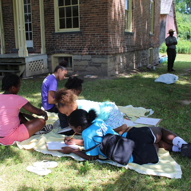 Several children in colorful clothing lay on a blanket in the grass. Behind them, a brick building.