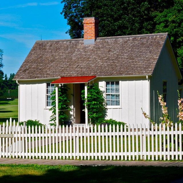 An open vista frames a white cottage with a gravesite and a flag in the distance.