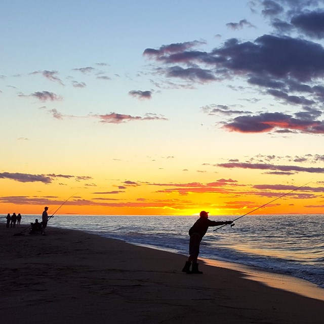 A bright sun sets behind the ocean as silhouettes of fishermen cast their lines out to sea.