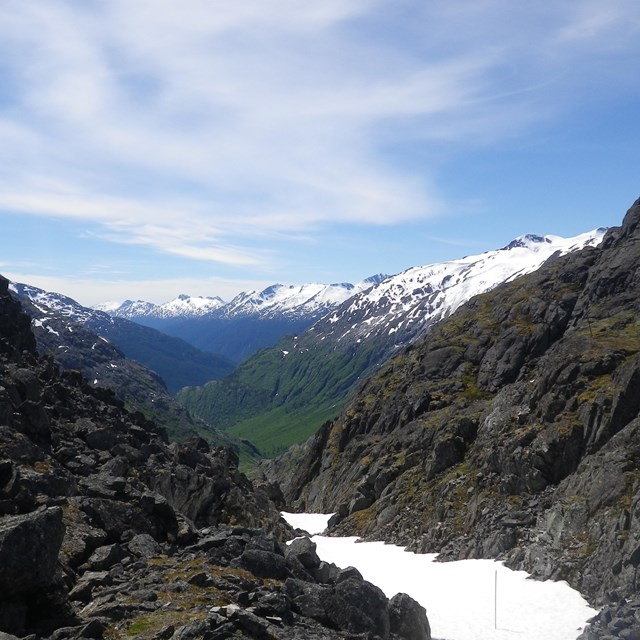 Looking from a rocky mountain valley with snow toward mountains and blue sky.