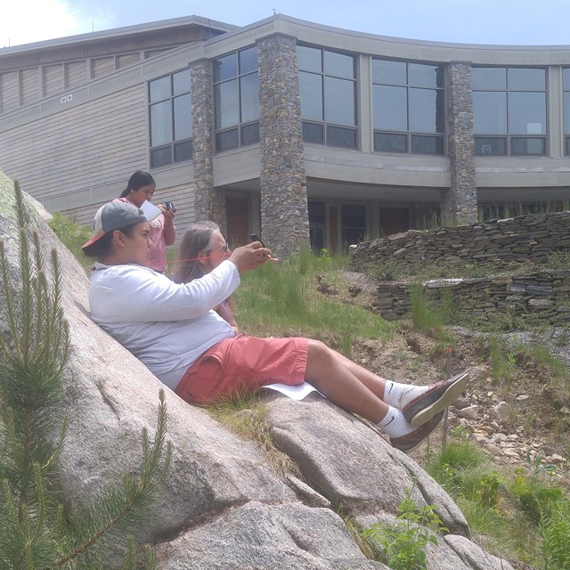 Tribal members sitting on large stones near a building and a grassy area.