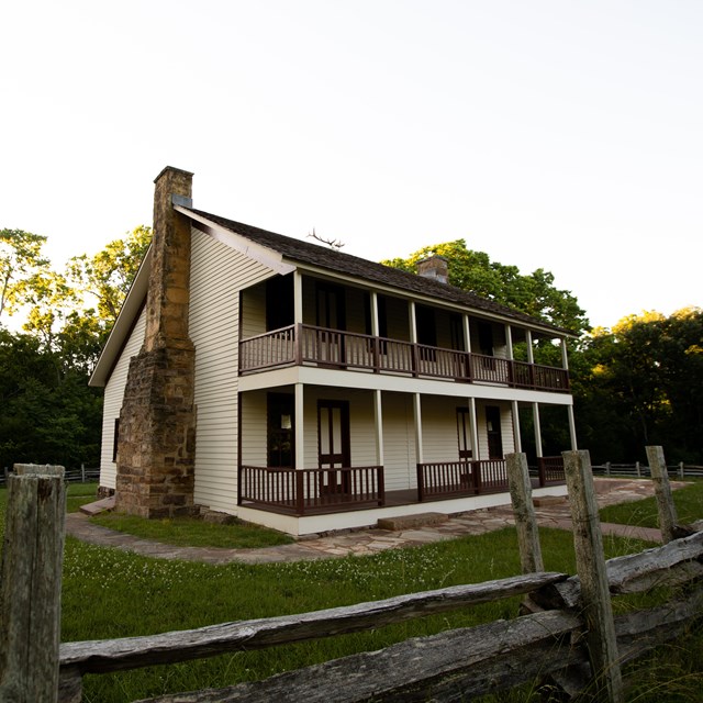 Two-story framed cream-colored house, two stone chimneys, porches with brown railing on both floors.