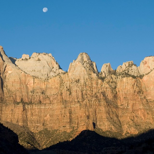 Large tan cliffs dotted with vegetation, blue sky with a moon above, and shadows in the foreground. 