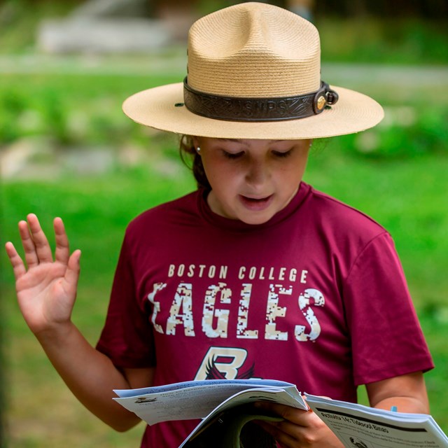 Two children in ranger hats speak while holding booklets, one with their hand raised