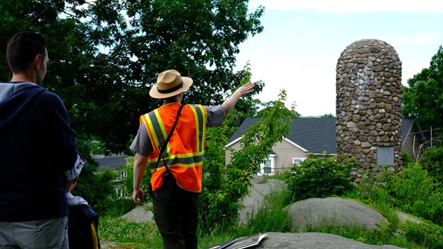 A park ranger wearing a safety vest points to a rock tower on top of a hill during a ranger talk