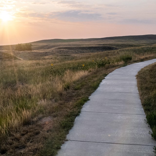 Concrete path curves uphill to right. Rolling hills and orange pink sunrise to left.