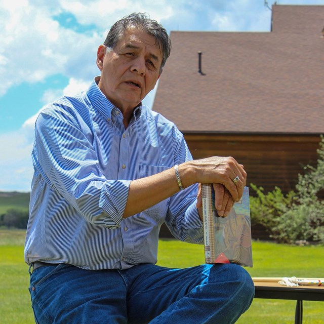 Man in light blue shirt and jeans sits on a stool holding a book against his knees, outdoors