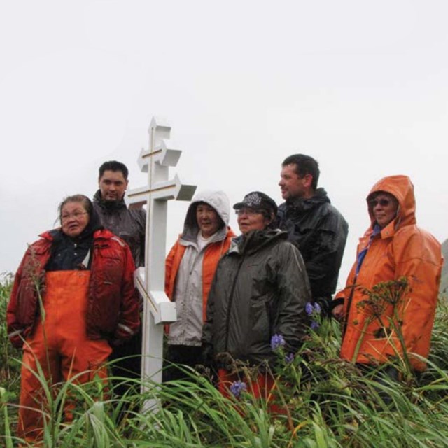 a group of people wearing orange rain slickers stand beside a white cross in a grassy field.