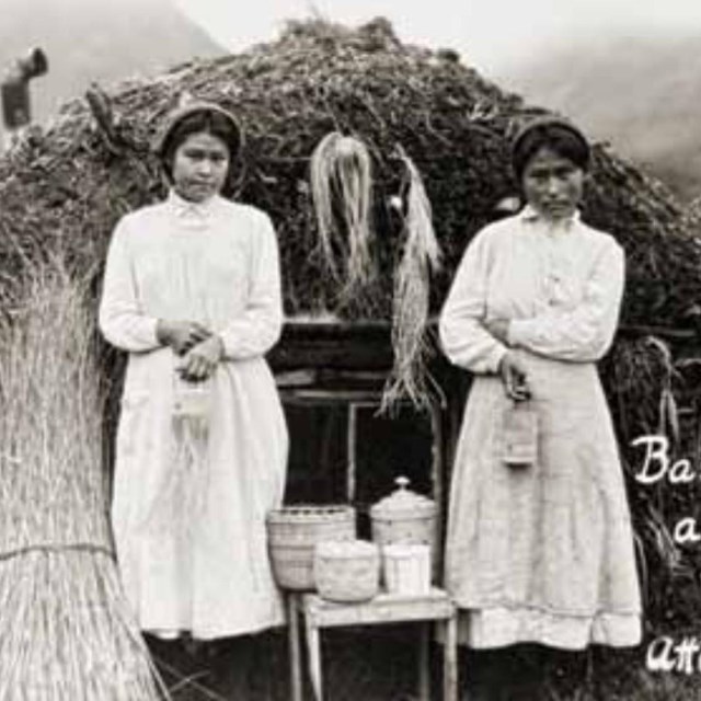 a historic scene of two women standing outside a sod house with fine grass baskets.