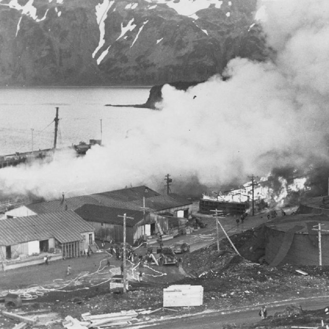 Black and white photo of a town with burning buildings and billowing smoke. 