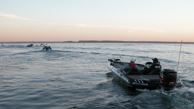 People in a motorboat with fishing gear on the lake at dawn with several other boats far ahead.