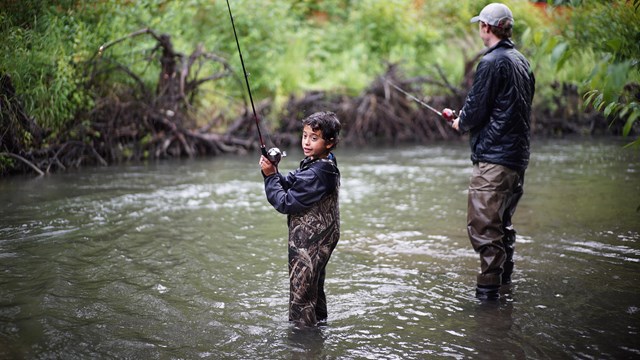 Outdoors; an adult and a child fishing in a stream - waders on and greenery on bank opposite them..