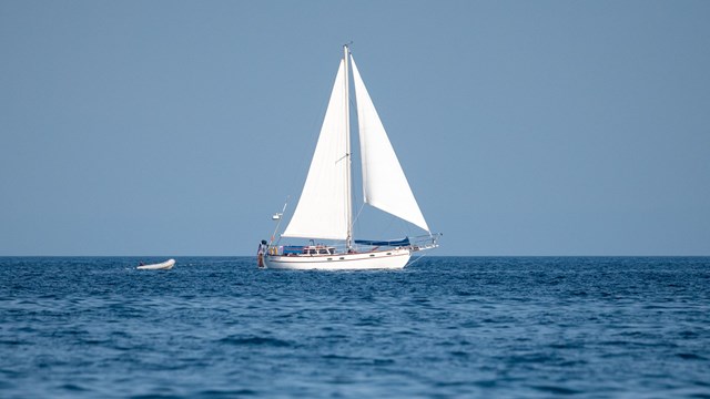 Sailboat with sails down resting on calm water with the sky and clouds reflecting off the surface. 